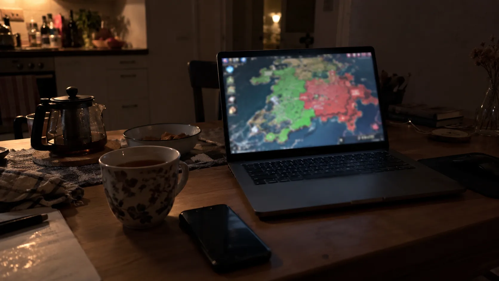 A laptop on a kitchen table late at night showing a blurred strategy game map, next to a cup of tea